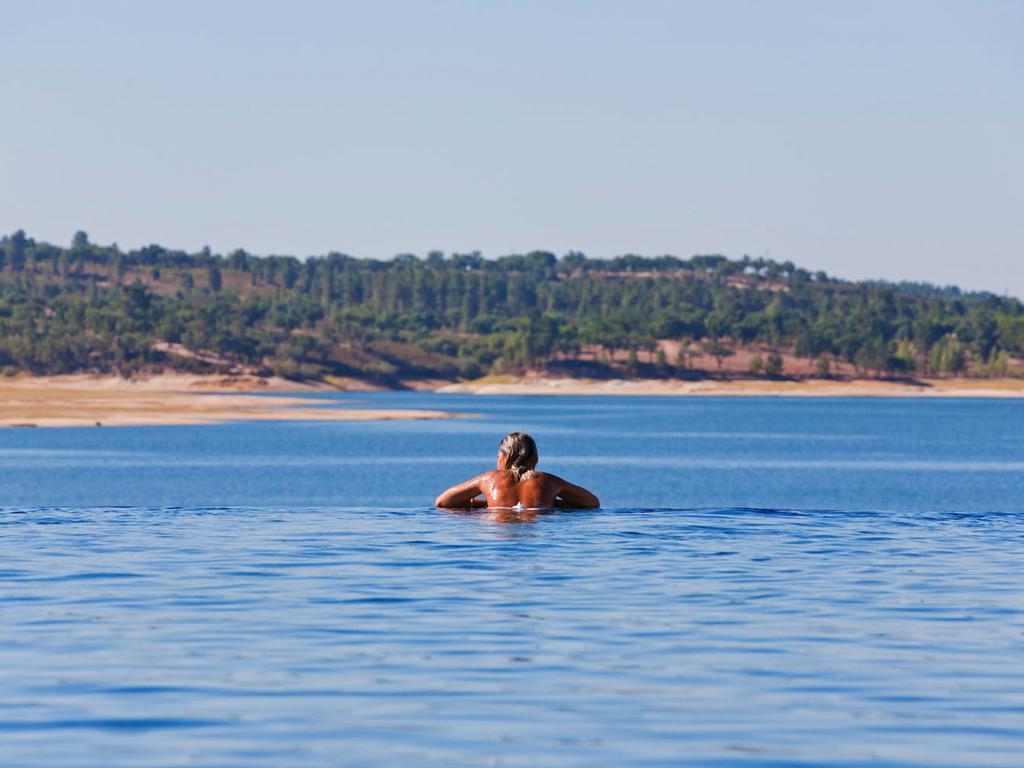 Ap Lago Montargil - Conference & Spa Exterior foto The photo shows a person swimming in a calm body of water, likely a lake or a river. The swimmer is seen from behind and appears to be enjoying the water on a sunny day. In the background, there's a scenic landscape featuring green trees and a sandy 