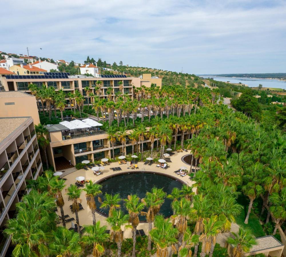 Ap Lago Montargil - Conference & Spa Exterior foto The photo shows a modern hotel complex set amidst lush greenery, featuring numerous palm trees. In the foreground, there is a circular swimming pool surrounded by lounge chairs. The hotel itself has multiple levels with balconies overlooking the pool