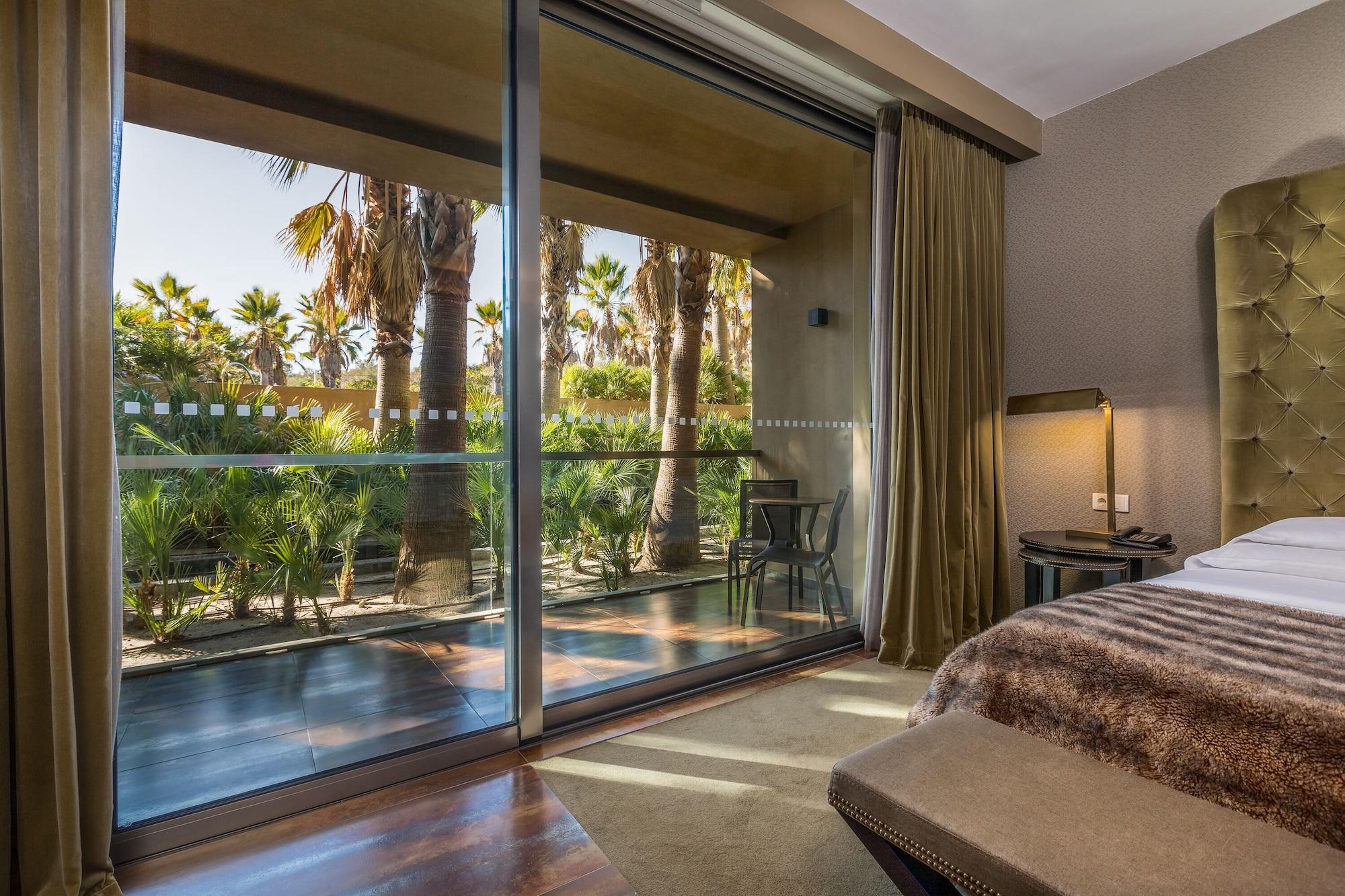 Ap Lago Montargil - Conference & Spa Exterior foto The photo shows a bedroom with large glass doors that lead out to a balcony or terrace. The view outside features palm trees and lush greenery, indicating a tropical or warm climate. Inside the room, there is a bed, a bedside table with a lamp, and a