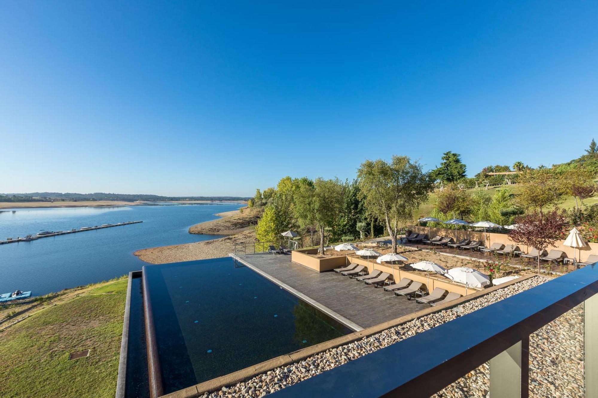 Ap Lago Montargil - Conference & Spa Exterior foto The photograph depicts a scenic view of a tranquil water body, likely a lake or river, bordered by lush greenery. In the foreground, there is an infinity pool that blends seamlessly with the water beyond. Comfortable lounge chairs are arranged on a w
